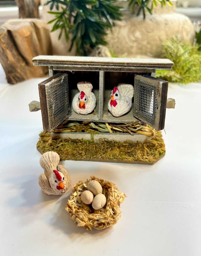 Puede incluir: Un gallinero de madera en miniatura con dos gallinas blancas dentro y un nido con tres huevos blancos fuera. El gallinero tiene una puerta de madera marr&oacute;n y una ventana de malla met&aacute;lica.
