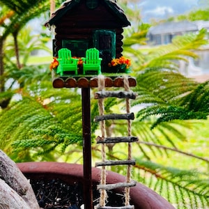 May include: A miniature wooden cabin with a green roof, a chimney, and a small porch with two green chairs. A wooden ladder made of twigs and twine leads up to the cabin. The cabin is set in a potted plant, surrounded by green foliage.