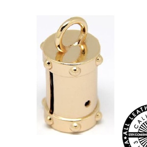 May include: Gold metal barrel-shaped charm with a loop at the top. The charm has a decorative design with small round studs around the top and bottom. The charm is made in the USA by All Leather Supplies, California since 2013.