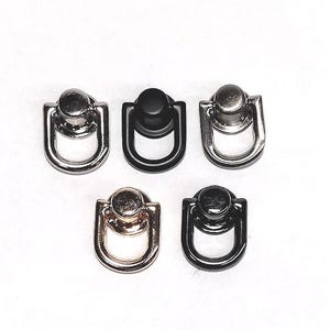 May include: Five metal D-ring connectors with a swivel base, arranged in a group. The set includes one silver, one matte black, one gunmetal, one rose gold, and one glossy black finish. Each piece features a circular stud and a D-shaped loop.