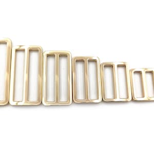 以下が含まれることがあります： さまざまなサイズの金色の長方形のスライダーバックルのコレクション。各バックルは3つのスロット設計で、バッグ、ベルト、その他のアクセサリーのストラップを調整するのに最適です。バックルは大きいものから小さいものへと配置されています。