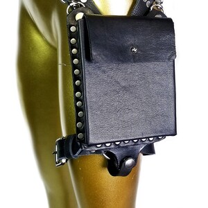 May include: Black leather thigh holster with a silver chain and metal accents. The holster has a flap with a snap closure and a small pocket on the front. The holster is decorated with silver studs.