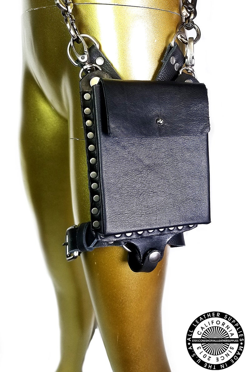 May include: Black leather thigh holster with a silver chain and metal accents. The holster has a flap with a snap closure and a small pocket on the front. The holster is decorated with silver studs.