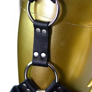 May include: Black leather harness with two silver metal rings and multiple silver metal studs. The harness is on a gold-colored mannequin.