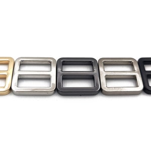 May include: Five metal rectangle slider buckles in a row. The buckles are in gold, silver, dark gray, light silver, and black. Each buckle has two rectangular openings. These are used for straps and belts.