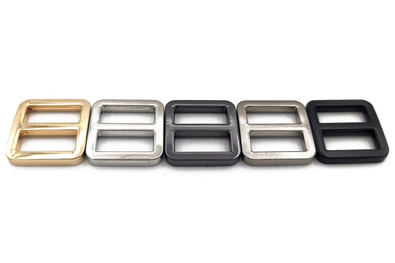 May include: Five metal rectangle slider buckles in a row. The buckles are in gold, silver, dark gray, light silver, and black. Each buckle has two rectangular openings. These are used for straps and belts.