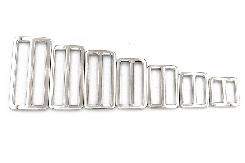 May include: A collection of seven shiny, silver-colored rectangular slider buckles, each with two vertical bars. The buckles vary in size, arranged from largest to smallest, and are suitable for various crafting and sewing projects.