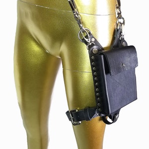 May include: Black leather thigh holster with a small pocket and a chain connecting to a belt. The holster is decorated with silver studs and has a buckle closure.