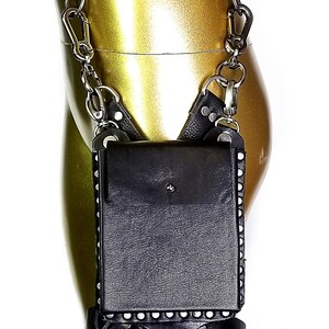May include: A black leather thigh holster with a silver chain and a silver snap closure. The holster has a small pocket on the front and is decorated with silver studs. The holster is attached to a brown leather belt.