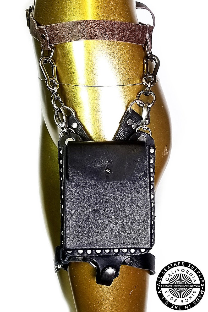 May include: A black leather thigh holster with a silver chain and a silver snap closure. The holster has a small pocket on the front and is decorated with silver studs. The holster is attached to a brown leather belt.