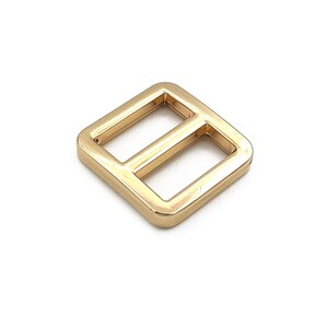 May include: A gold-colored metal slider buckle with a square shape and two rectangular openings. The hardware is likely used for straps or belts. The buckle is shown against a white background.
