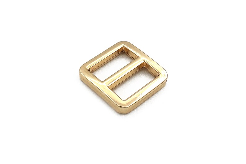 May include: A gold-colored metal slider buckle with a square shape and two rectangular openings. The hardware is likely used for straps or belts. The buckle is shown against a white background.