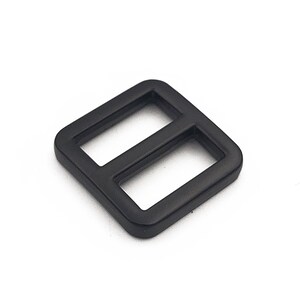 May include: A black, square-shaped slider buckle with two rectangular openings. The buckle is made of a matte material and is set against a white background. This hardware component is likely used in bags, straps, or other accessories.