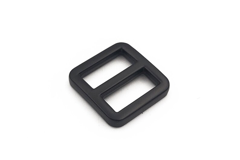 May include: A black, square-shaped slider buckle with two rectangular openings. The buckle is made of a matte material and is set against a white background. This hardware component is likely used in bags, straps, or other accessories.