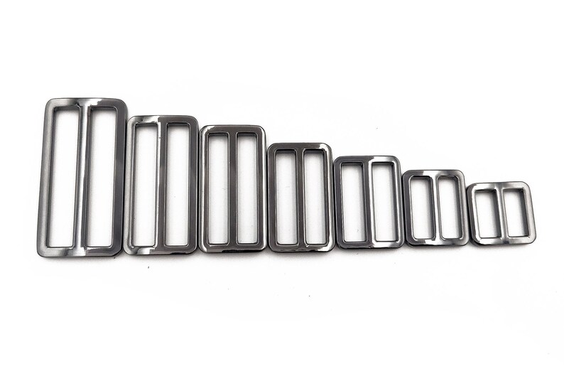 May include: A collection of seven silver-colored rectangular metal slides, each with two interior bars. The slides are arranged in descending order by size, from largest to smallest, against a white background. These are likely used for straps or belts.