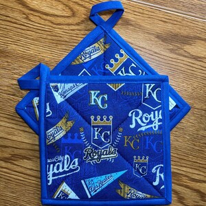 May include: Two blue quilted pot holders with a Kansas City Royals theme. The fabric features the team's logo, crowns, and text in white and gold. Each holder has a blue loop for hanging.