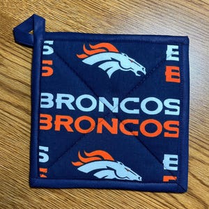 May include: A square navy blue quilted pot holder featuring a repeating pattern of the Denver Broncos horse logo in white and orange, along with the text BRONCOS printed in white and orange block letters, complete with a small corner hanging loop.