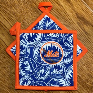 May include: A square quilted pot holder featuring a blue fabric with a repeating white pattern of baseballs and the New York Mets logo. The center displays a circular patch with the Mets logo and a city skyline. It has an orange fabric border.