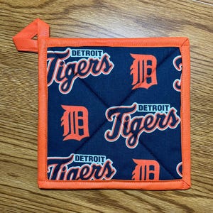 May include: A square Detroit Tigers pot holder with a navy blue background and orange trim. The pot holder features the team's logo and the words "Detroit Tigers" in orange and white.