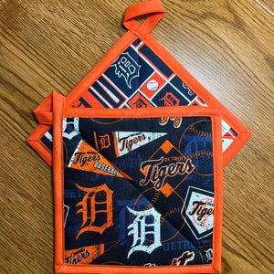 May include: A square fabric pot holder featuring a dark blue background with a repeating Detroit Tigers baseball pattern in orange and white. The design includes team logos, pennants, and baseballs, finished with a bright orange fabric border.