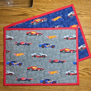 May include: Two fabric placemats with a red border. The top placemat is blue with white Hot Wheels logos and a repeating pattern of red, white, and blue race cars. The bottom placemat is gray with white Hot Wheels logos and a repeating pattern of red, white, and black race cars.