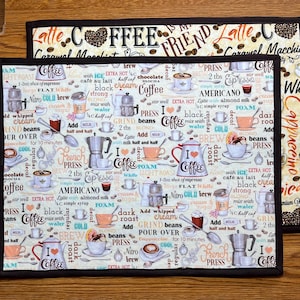 May include: A beige fabric placemat with a black border features a coffee-themed design. The placemat is covered in coffee-related words and illustrations, including coffee beans, coffee cups, and coffee makers. The text on the placemat includes "Coffee is my friend", "Caramel Macchiato", "Americano", "Espresso", "Cappuccino", "Nitro Cold Brew", "French Press", "Grind Beans", "Pour Over", "Extra Hot", "Light Roast", "Dark Roast", and "I Love Coffee".