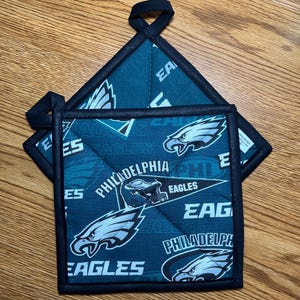 May include: Two Philadelphia Eagles themed pot holders. The pot holders are teal with a black border and a repeating pattern of the team's logo and the words "Eagles" and "Philadelphia". Each pot holder has a black loop for hanging.