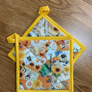 May include: Two yellow quilted pot holders with a floral and bee print. The print features sunflowers, daisies, honeycombs, and bees. The text on the print includes "Sold by the Pound" and "Our Own Honey".