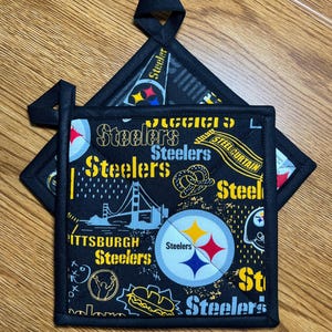 May include: Two square fabric pot holders featuring a black background with a repeating Pittsburgh Steelers pattern in yellow, white, blue, and black. The design includes the team logo, the word Steelers, a bridge, a football, and a pretzel.