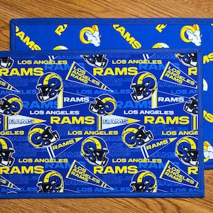 May include: Two rectangular placemats with a blue background and a repeating pattern of yellow and black Los Angeles Rams football helmets, the word "RAMS", and the team logo. The placemats have a blue border.