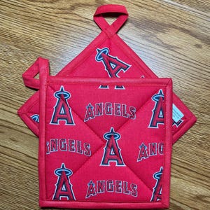 May include: Red quilted pot holders featuring the Los Angeles Angels baseball team logo and wordmark. The pot holders have a square shape with a loop for hanging. The design is repeated across the fabric.