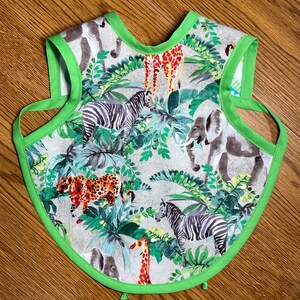May include: A green and white bib with a jungle animal print. The bib features zebras, giraffes, elephants, and leopards. The bib has a green trim around the edges.