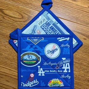 May include: Blue and white quilted pot holders featuring Los Angeles Dodgers logos and text. The pot holders have a loop for hanging and include the text "Dodgers", "Los Angeles", and "Vin Scully Ave".