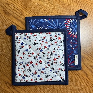 May include: Two blue and white quilted pot holders with a red, white, and blue star pattern and a red, white, and blue firework pattern. The pot holders have a blue border and a loop for hanging. The pot holders are handmade by "The Handmade Soap Lady".
