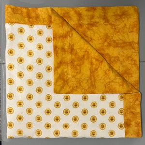 May include: A white fabric with a pattern of yellow smiling suns is folded in half with a solid yellow fabric on top. The yellow fabric has a mottled, tie-dye effect.