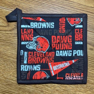May include: A square black pot holder with a loop, featuring a repeating pattern of orange and white text and football helmets. The text includes "BROWNS", "DAWG POUND", and "CLEVELAND".