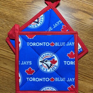 May include: Two blue and red Toronto Blue Jays pot holders. The pot holders feature a repeating pattern of the team's logo and the words "Toronto Blue Jays". Each pot holder has a red border and a loop for hanging.