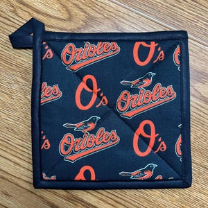 May include: Black square pot holder with a navy blue border and loop. The pot holder features a repeating pattern of the Baltimore Orioles logo and bird in orange and white. The pot holder is quilted.