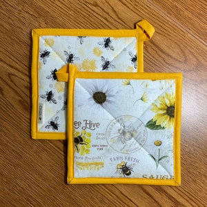 May include: Two yellow and white quilted pot holders with a beehive and sunflower design. The text on the pot holders reads "Bee Hive", "Your Name's", "Open Daily", "Unfiltered Pure", "Spring Products", "South Carolina", "Welcome", "Farm Fresh", and "Sauce".