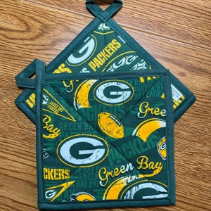 May include: Two green and yellow quilted pot holders with a Green Bay Packers theme. The pot holders feature the team's logo, footballs, and the words "Packers" and "Green Bay". Each pot holder has a loop for hanging.