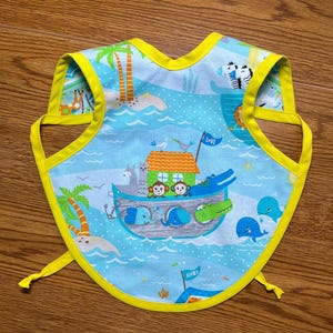 May include: A baby bib with a blue background and a yellow trim. The bib features a colorful illustration of Noah's Ark with various animals, palm trees, and the word "LOVE" on a flag. The bib has ties on each side.