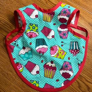 May include: A turquoise blue bib with a red trim and a pattern of colorful cupcakes. The cupcakes have pink, yellow, green, and blue frosting and are decorated with cherries and sprinkles.