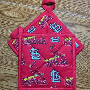 May include: Red pot holders with a quilted pattern featuring the St. Louis Cardinals logo, a cardinal bird, and the word "Cardinals" in a script font. The pot holders have a loop for hanging.