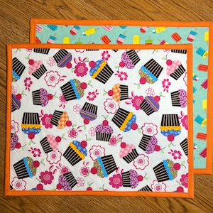 May include: Two fabric placemats with orange trim. The top placemat has a light blue background with colorful popsicle designs. The bottom placemat has a white background with a repeating pattern of colorful cupcakes.