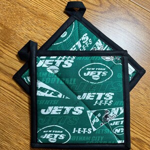 May include: Two square quilted pot holders featuring a green fabric with a white New York Jets football team print. The pattern includes the team name, logos, and the text J-E-T-S. Each pot holder has a black fabric border and a small hanging loop.