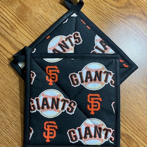 May include: Black quilted pot holders with a baseball and the text "GIANTS" in orange and white. The pot holders have a loop for hanging and are made of a durable fabric. The design features the San Francisco Giants logo.