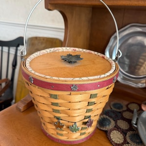 Puede incluir: Una cesta tejida redonda con tapa de madera que presenta un asa de metal en forma de hoja de arce. La cesta tiene un borde rojo, un asa metálica abatible con agarre de madera y detalles decorativos de hojas de arce en metal.