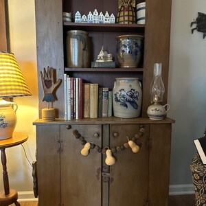 May include: A rustic wooden cabinet with open shelves displaying various antique items. The shelves hold stoneware crocks, books, and decorative figurines. A string of gourds and pinecones hangs from the cabinet doors. A lamp with a yellow lampshade is visible on the left.