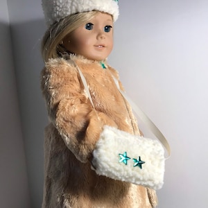 Puede incluir: Una muñeca con un abrigo de piel sintética blanca y un gorro de piel sintética blanca con estrellas turquesa. La muñeca lleva un bolso de piel sintética blanca con estrellas turquesa.