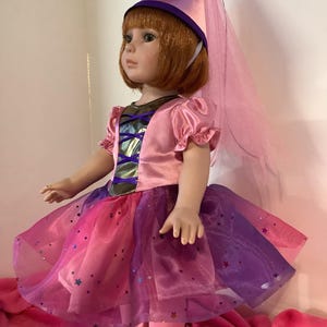 May include: A pink and purple fairy costume for a doll, featuring a tulle skirt with glitter stars, a pink satin bodice with a gold trim, and a pink cone hat with a purple trim and a tulle veil.
